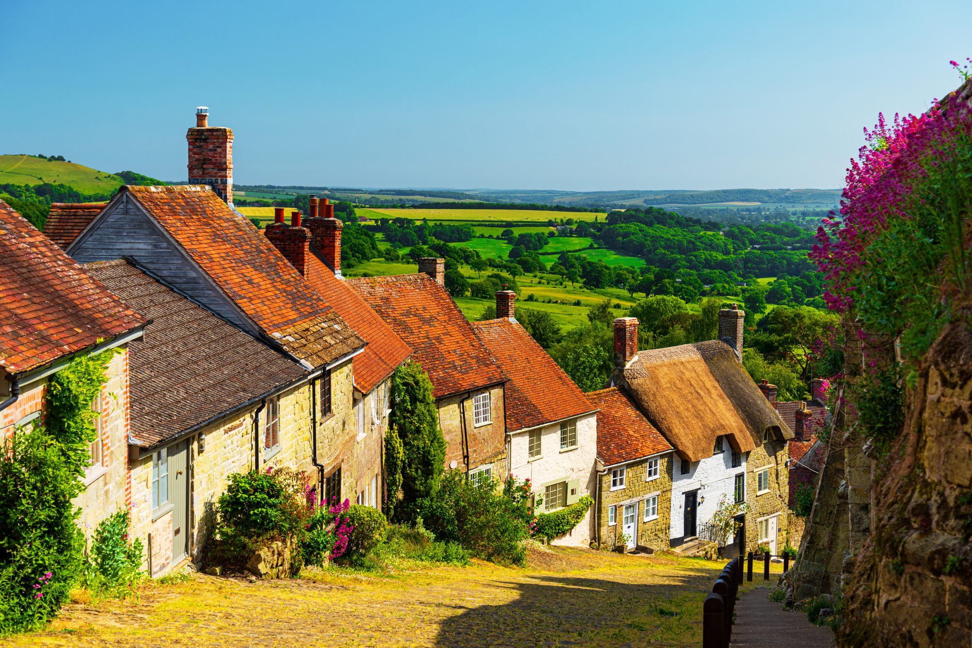 View down the historic cobbled Gold Hill in Shaftesbury, Dorset, with stone cottages lining the steep street and rolling countryside beyond.